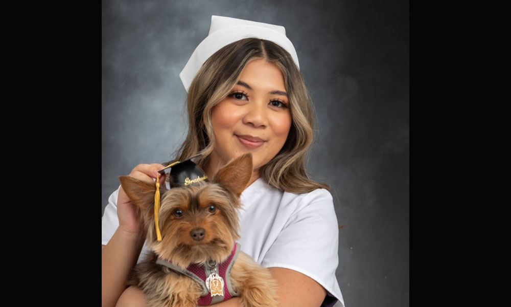 Nicole poses in a nursing uniform holding her dog who is wearing a graduation cap