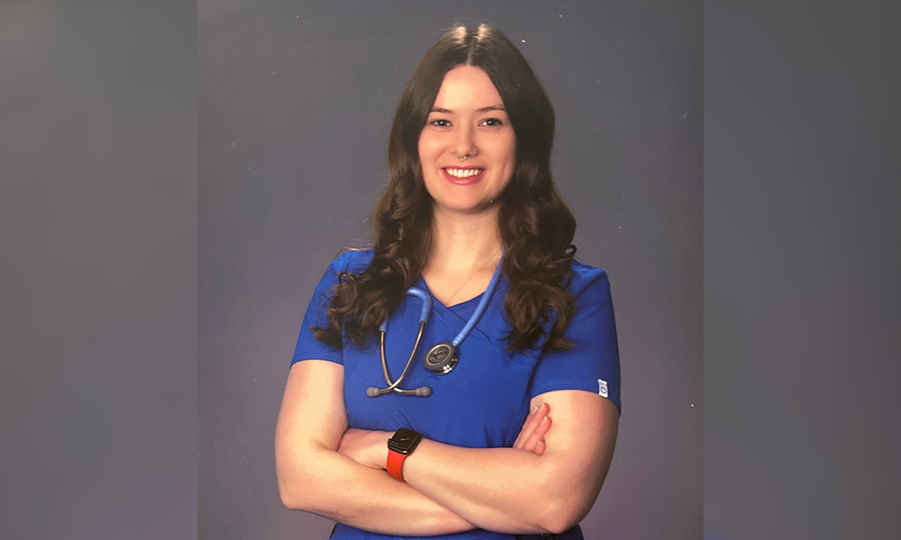 Shelby is smiling at the camera, wearing blue nursing scrubs, in front of a grey background