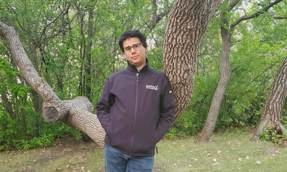 A man in glasses and a grey sweater leans against a tree.