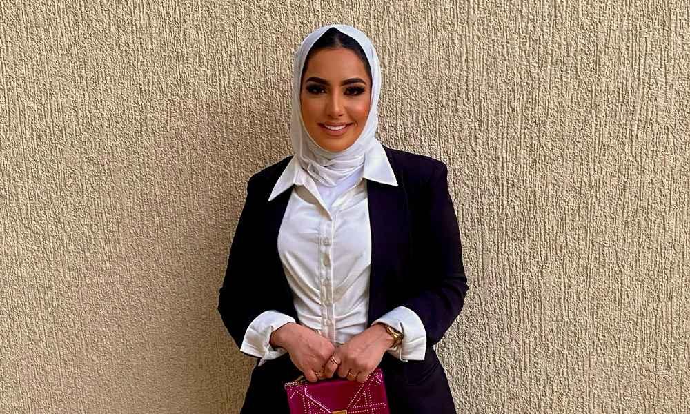 A woman in a white hijab and black blazer stands in front of a stucco wall.