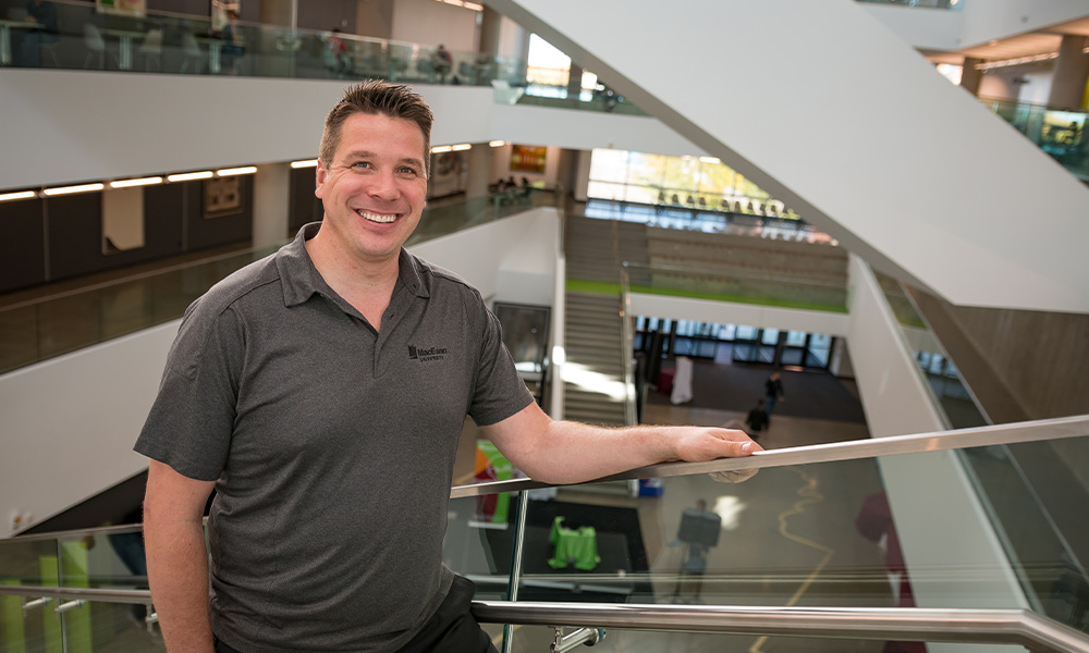 Dr. Mike Annett wears a grey MacEwan polo tee and leans against a glass railing in Allard Hall