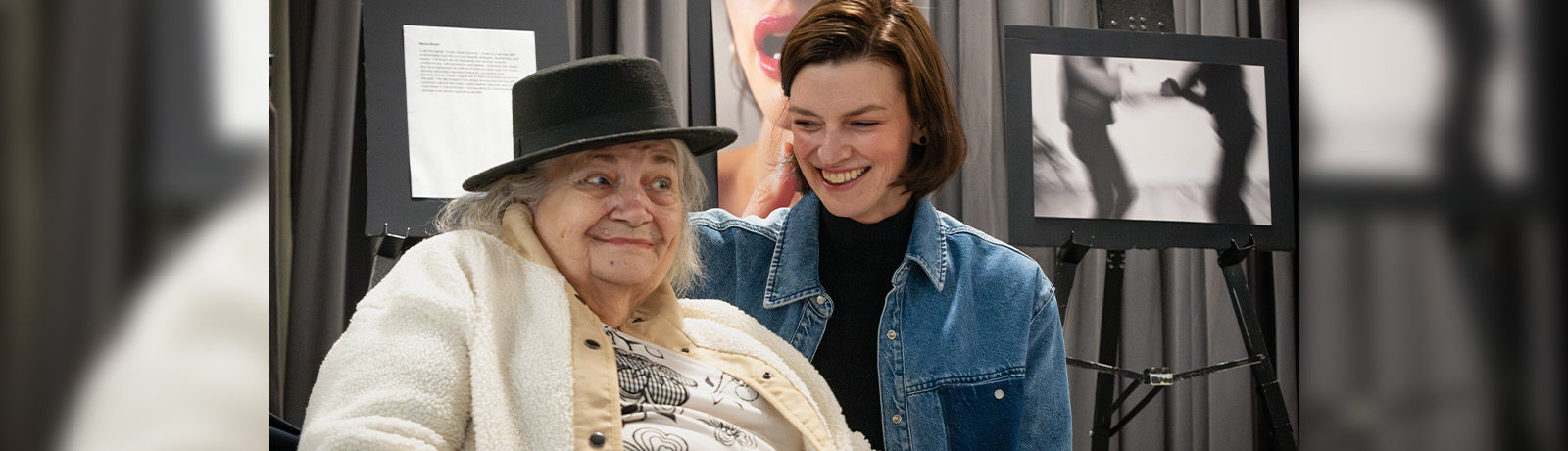 A senior woman wearing a hat sits with a young woman, both smiling.