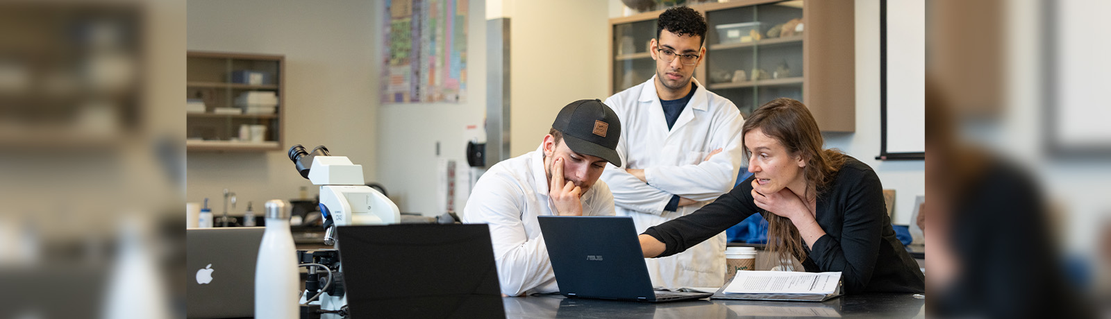 A woman points to something on a laptop screen while two male students in lab coats observe.