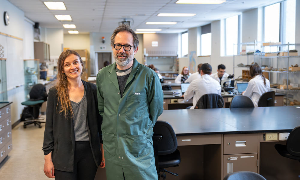 A man and a woman stand side-by-side in a lab with students working in the background.