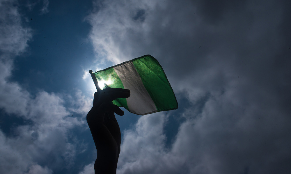 A hand holds a Nigerian flag against a background filled with sky