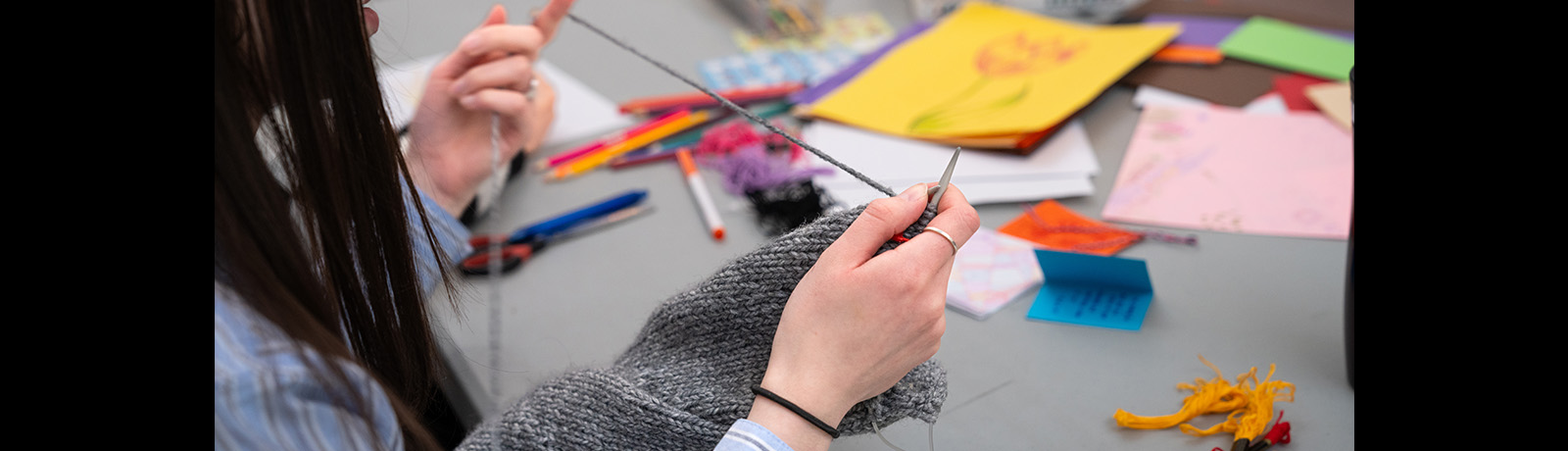 A student knitting against the backdrop of a table filled with art supplies