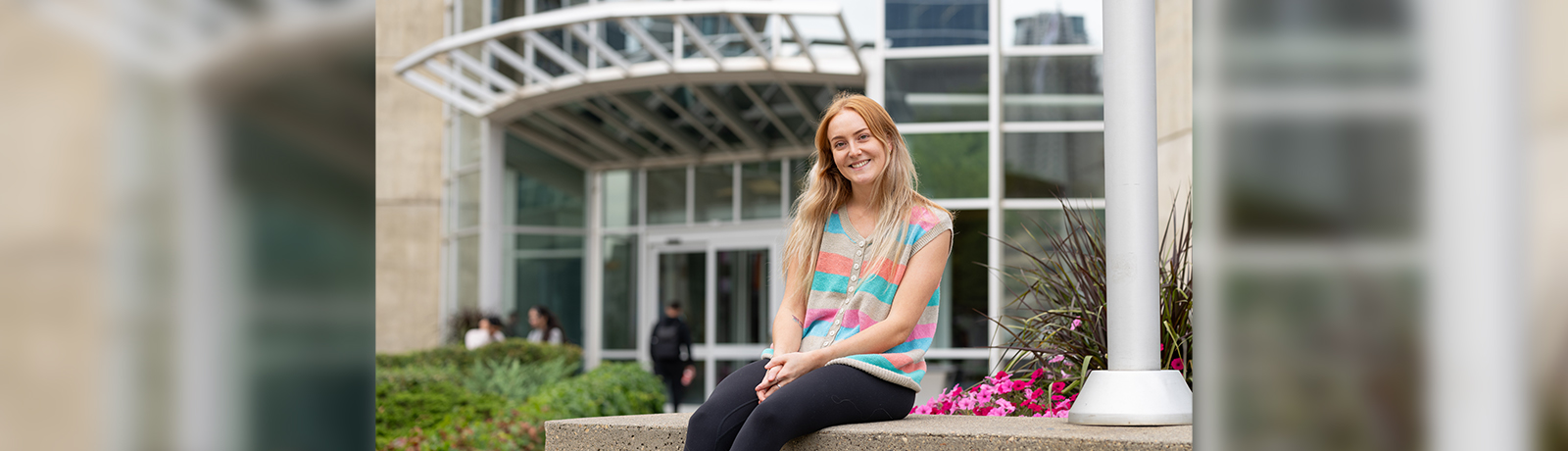 Dana Jones sits on a concrete block in front of a building, smiling, hands folded in her lap