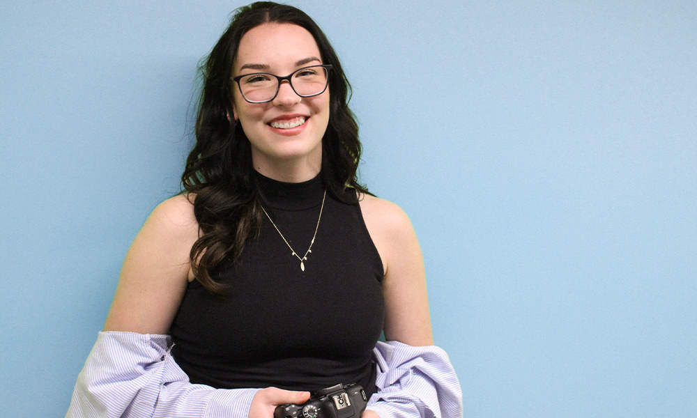 Myah Juneau stands in front of a blue backdrop, holding a camera.