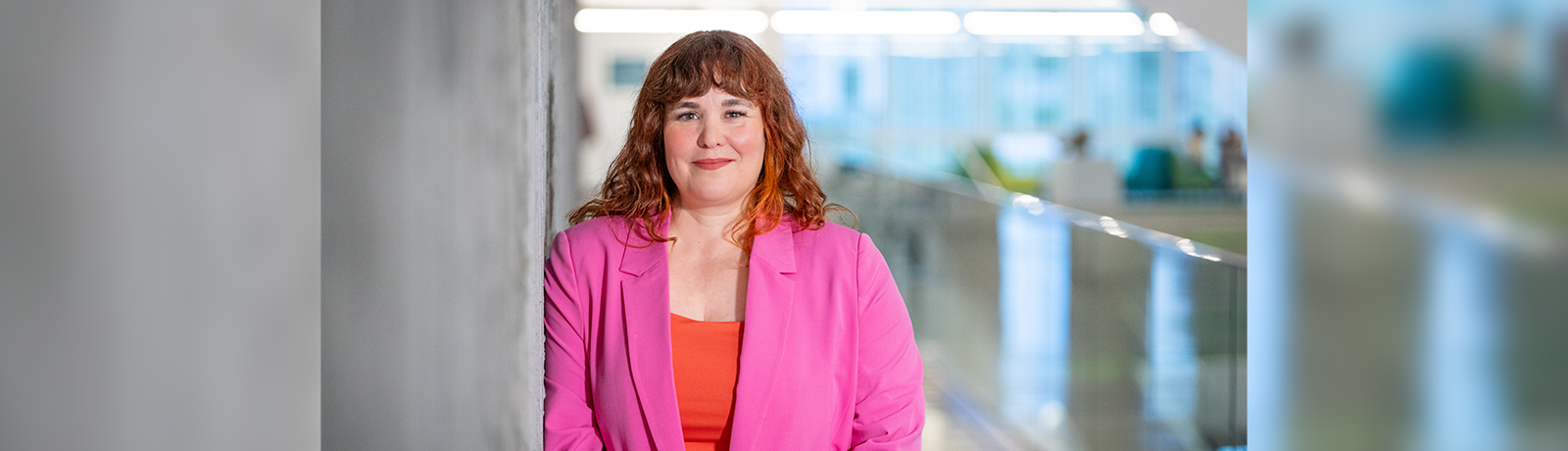 A woman stands against a concrete pillar, smiling, wearing a pink blazer and a orange shirt."