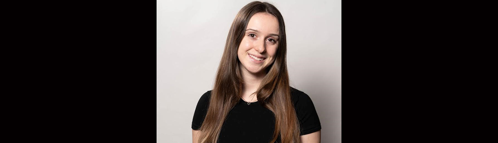 A woman in a black t-shirt stands smiling in front of a grey wall