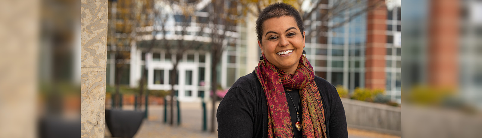 Jenna Butler wears a black shirt and a red patterned scarf, and sits in front of the clocktower entrance of Building 6.