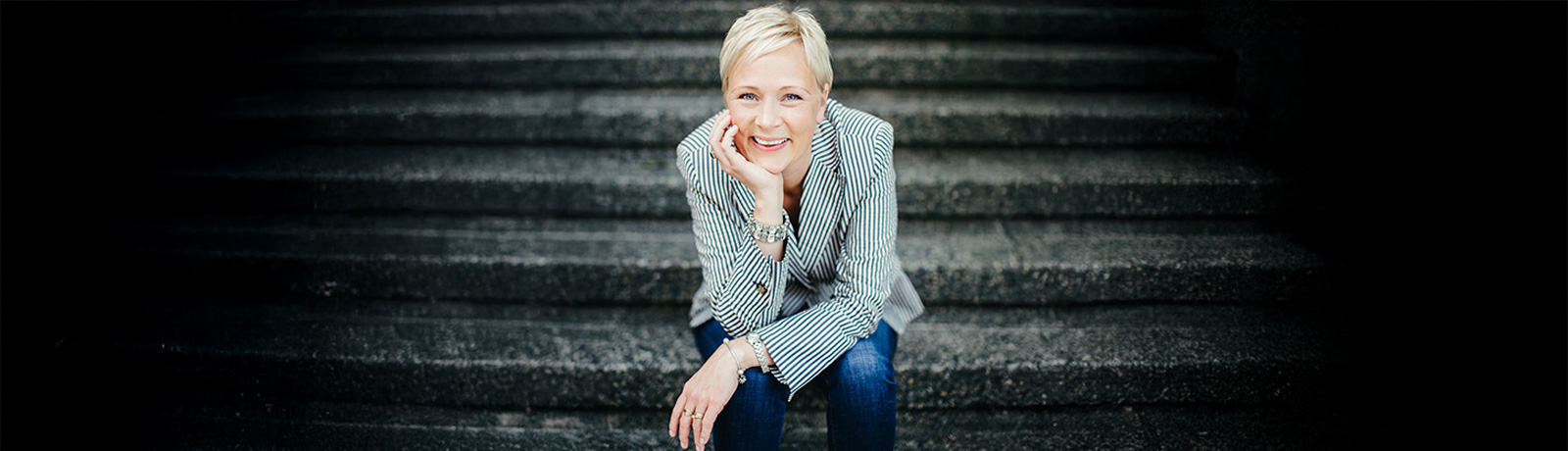 A woman smiling, sitting down and leaning forward with her head in her hand, her elbow on her knee