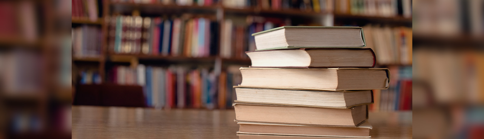 A stack of books sit on a table with a large bookshelf in the background.