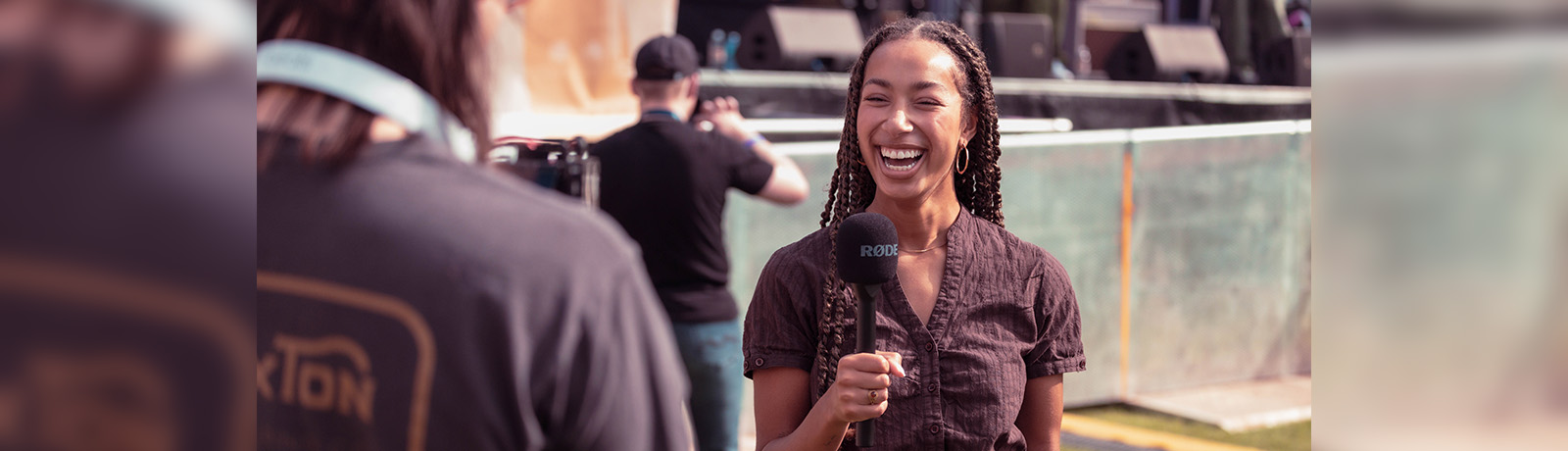 Aajah Sauter stands in front of a camera, holding a microphone and smiling.