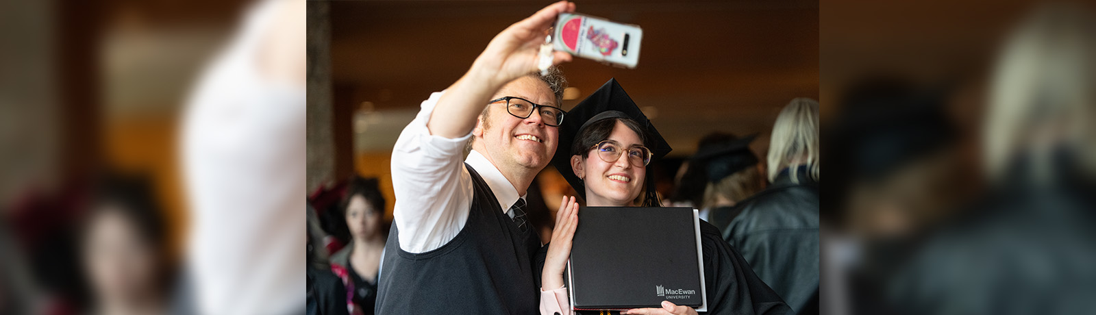 A man takes a selfie on a cell phone with a female grad in a cap and gown holding a degree.