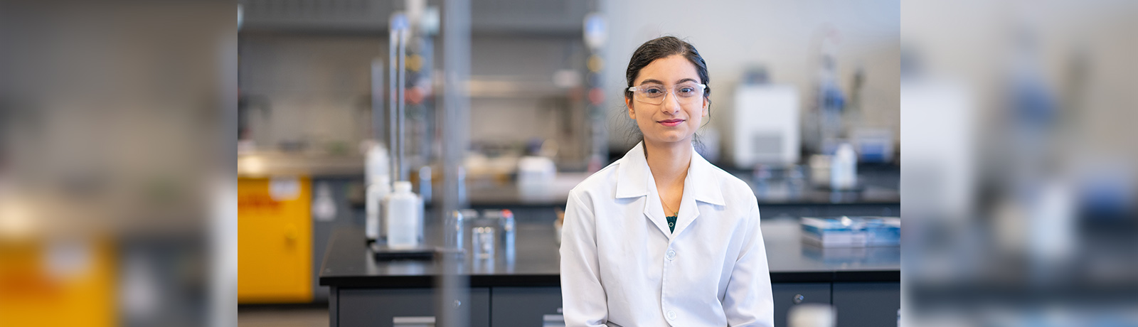 "A student sits at a lab table wearing a white lab coat and safety goggles