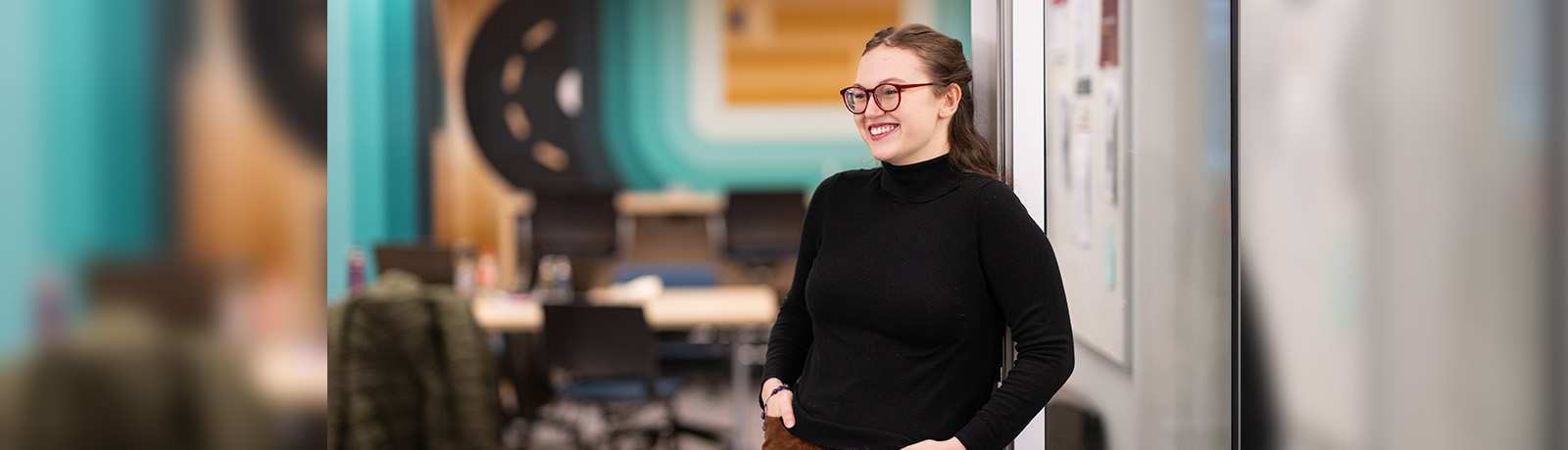 Beth Murray wears a black long-sleeved shirts and stands in the Writing Centre, hands in pockets, smiling at something off camera.