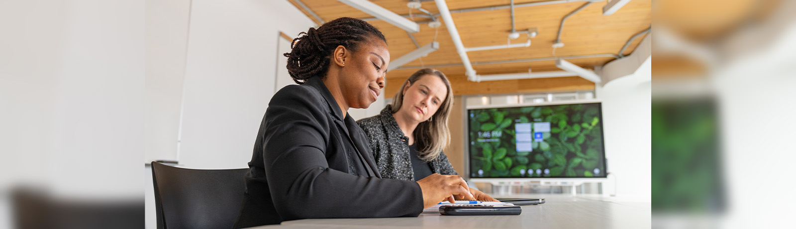 Two women sit at a conference table, looking down at a tablet screen.