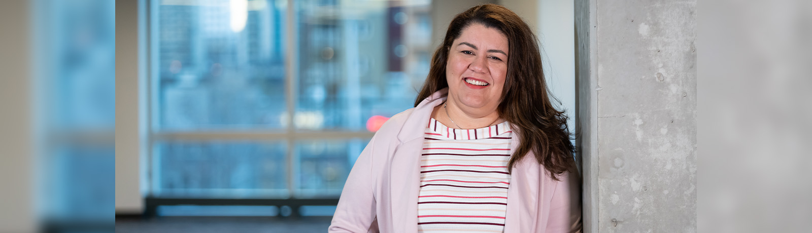 Dr. Fernanda Dos Santos leans against a concrete pillar, smiling, wearing a pink cardigan over a striped shirt.