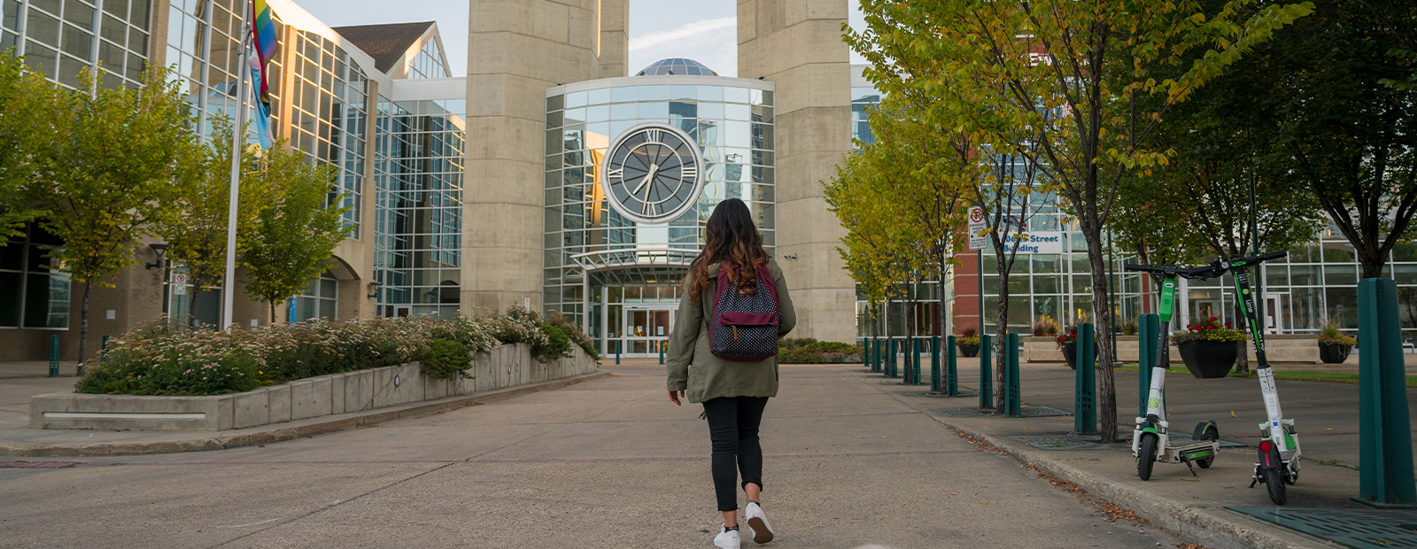 A woman with dark hair, wearing a green jacket and a backpack, is seen from behind as she walks toward the clock tower entrance on MacEwan's campus.
