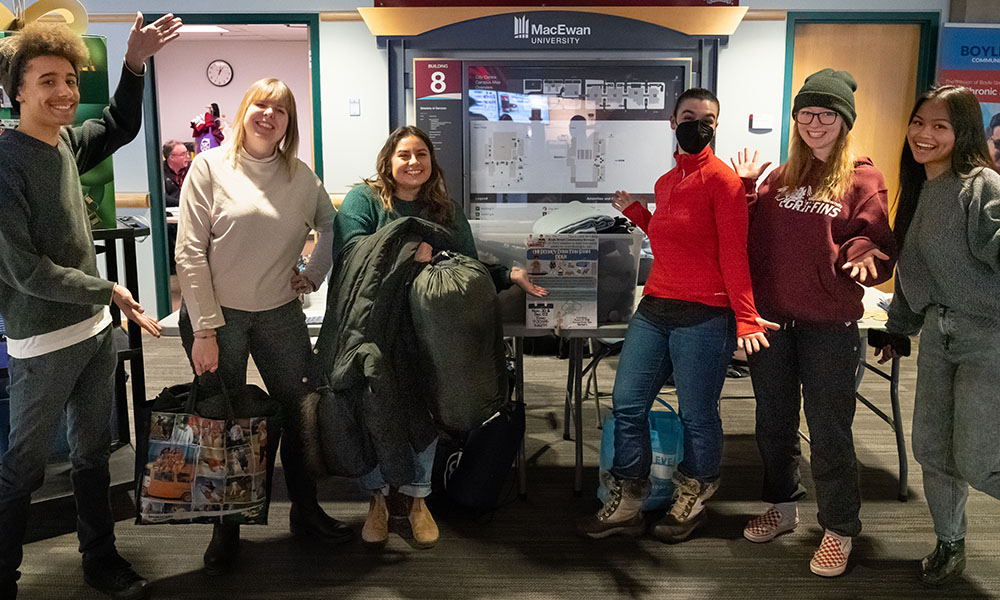 Students in INTD 205 stand in front of a table in Building 8 with bins to collect warm clothing